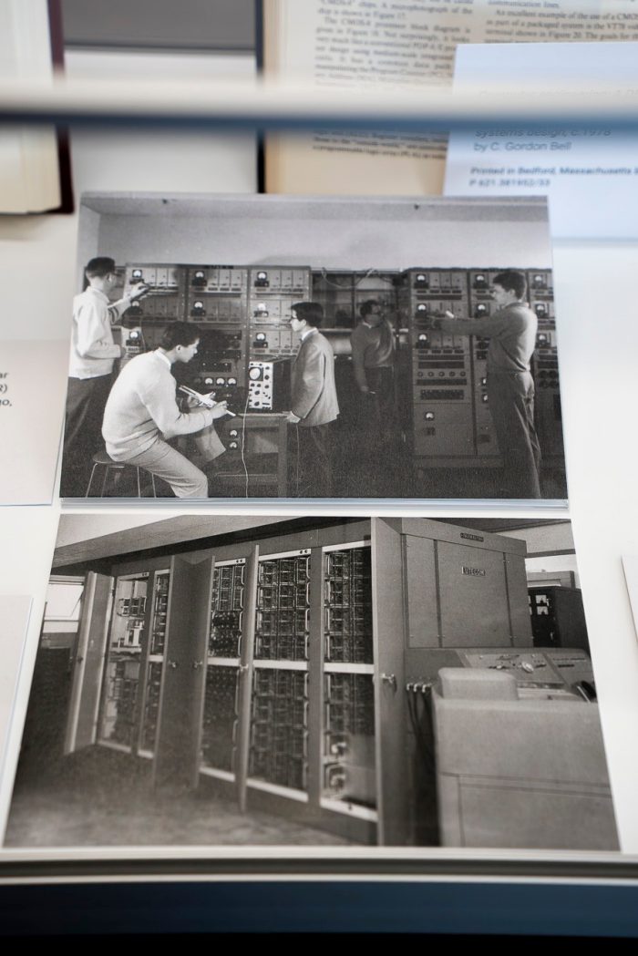 A close up view of two black and white photos on in a case display case. The photos show images of vintage 1950s computers. 