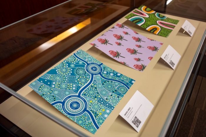 A side view of a display case with three colourful artworks inside. The closest artwork at the left of the photo shows an strikingly blue artwork with a series of lines and dots.