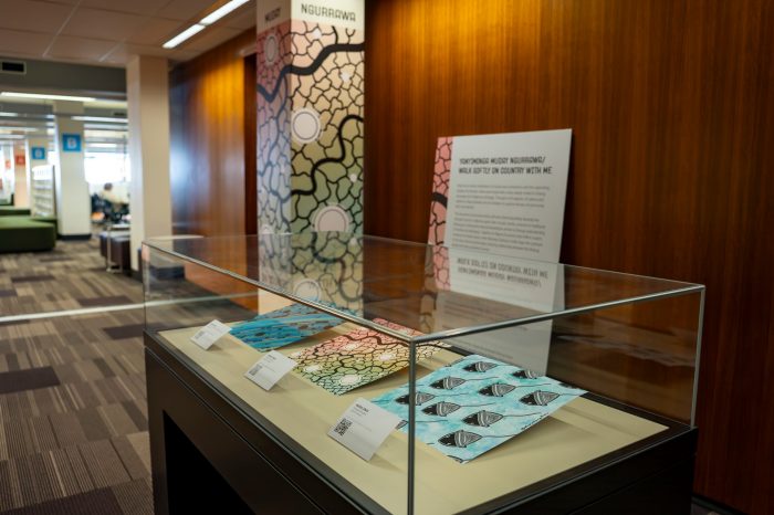 A side on view of a glass display case with three artworks inside. Behind the display case is a column covered in a colourful patterned print, similar to one of the artworks in the display case. 
