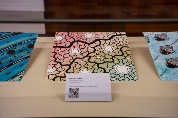 A close up on a digital illustration in a display case. The illustration shows fragmented black lines and white circles on top of a colour gradient moving from pink, to orange, to blue. 