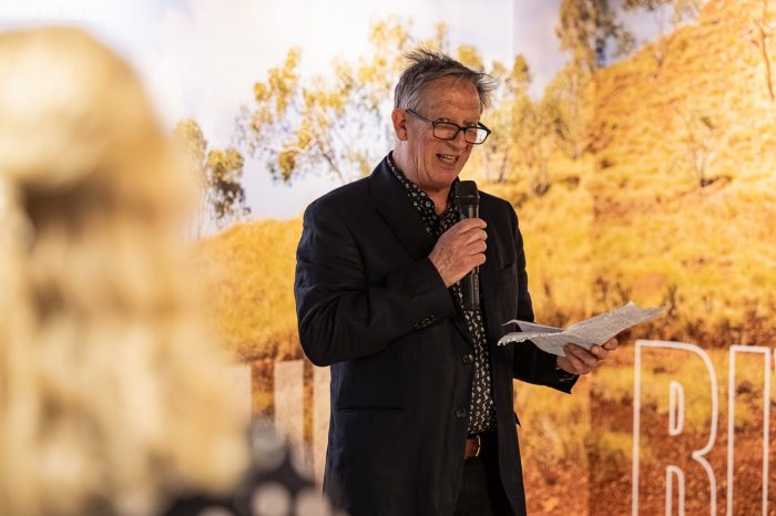 Revealing Riversleigh - Professor John Long at the opening night A man wearing a dark suit and glasses holds a microphone and addresses an audience.