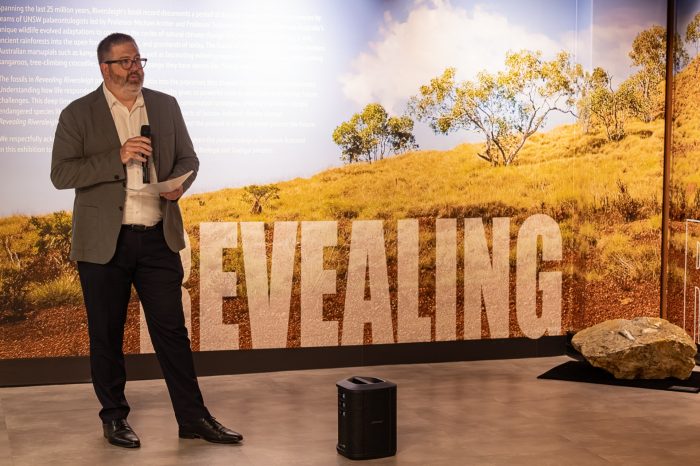 Revealing Riversleigh - Professor Dane McCamey, PVC Research UNSW, at the opening night A man in a grey suit holding a microphone addresses and audience. The title wall of the exhibition "Revealing Riversleigh" is in the background.