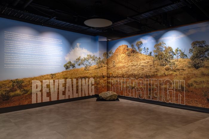 Revealing Riversleigh - installation view - title wall An entrance to a gallery space with a large image of a rocky, arid landscape imposed over two walls. Across the walls is the title text of the exhibition in large letters, "Revealing Riversleigh". In the middle of the two walls is a large rock on the floor.