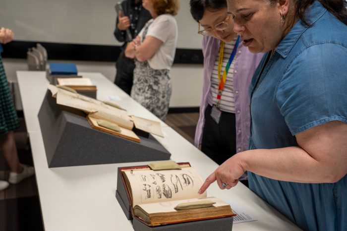 People stand and look at old books propped open on a table. A woman in blue points at the book.