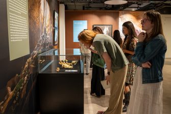 Students stand near a glass display case containing fossils. One student is leaning down to look at an item within.