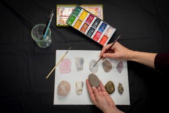 Close up of a table with watercolour paints and someone creating a colourful painting of different coloured rocks.