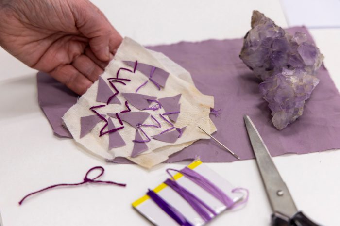 A person's hand holds a craft project of a small piece of fabric with purple triangles stitched onto it. Nearby are purple thread, scissors, a needle, and an amethyst crystal.