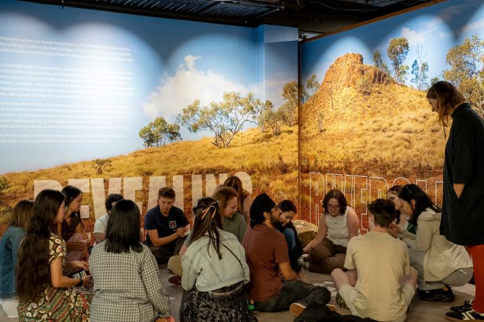 A group of university students sitting in a circle on the exhibition space floor in front of large, backlit murals depicting the dry Riversleigh landscape of northern Queensland.