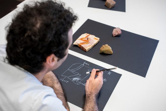 A person sketches geological diagrams with a white pencil on black paper while observing rock and mineral specimens.