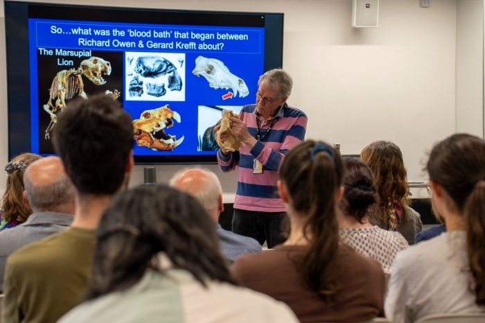 A man in a striped shirt holds a skull specimen while presenting to a seated audience. Behind him, a screen displays images of a marsupial lion skeleton and fossils.