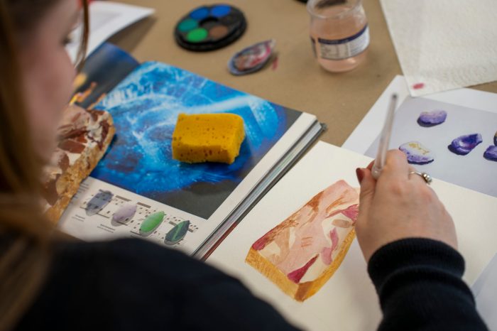 A person holding a paintbrush creating a watercolour illustration of a textured rock, referencing an image in an open book.