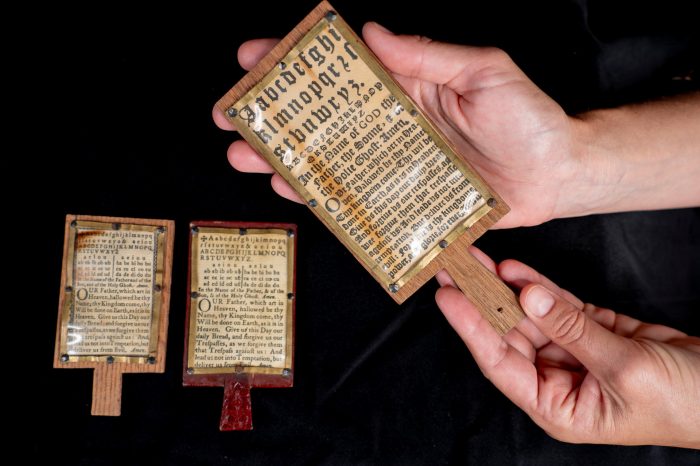 Three small wooden horn-books with handles, displaying the alphabet and Lord's Prayer, are held by a hand.