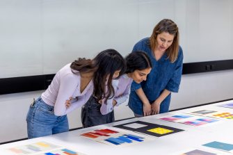 Three women lean over a white table looking at a series of prints depicting colourful shapes. 