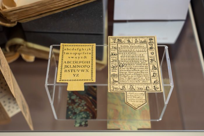 A close-up photo of two paddle-shaped cardboard horn books. The horn book on the left has the English alphabet typed on it, the right horn book has a series of small black and white images and text. 