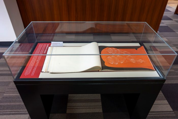A display case containing a single, large open book displaying a red and black illustration of the Sydney Opera House