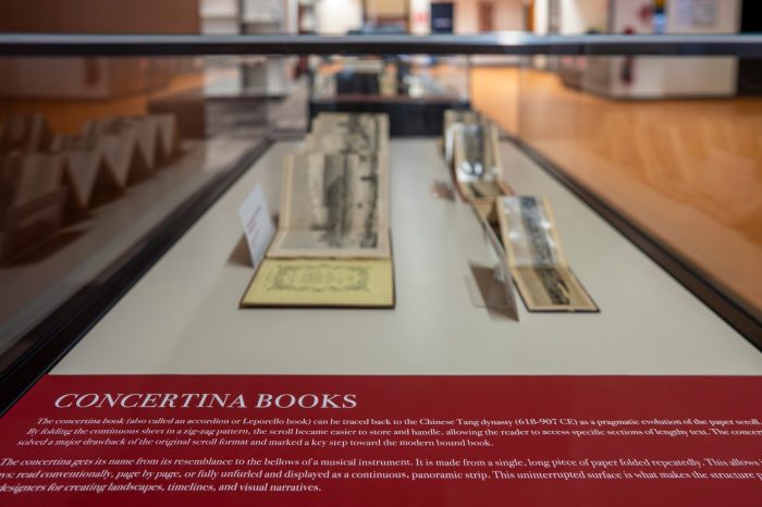 A close-up of a glass display case containing a red label that reads "Concertina Books". There are three opened concertina books in the background. 