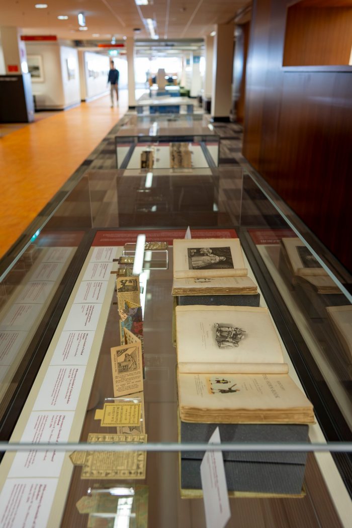 A view of four glass display cases in a row, with a person walking down a hallway in the background. 