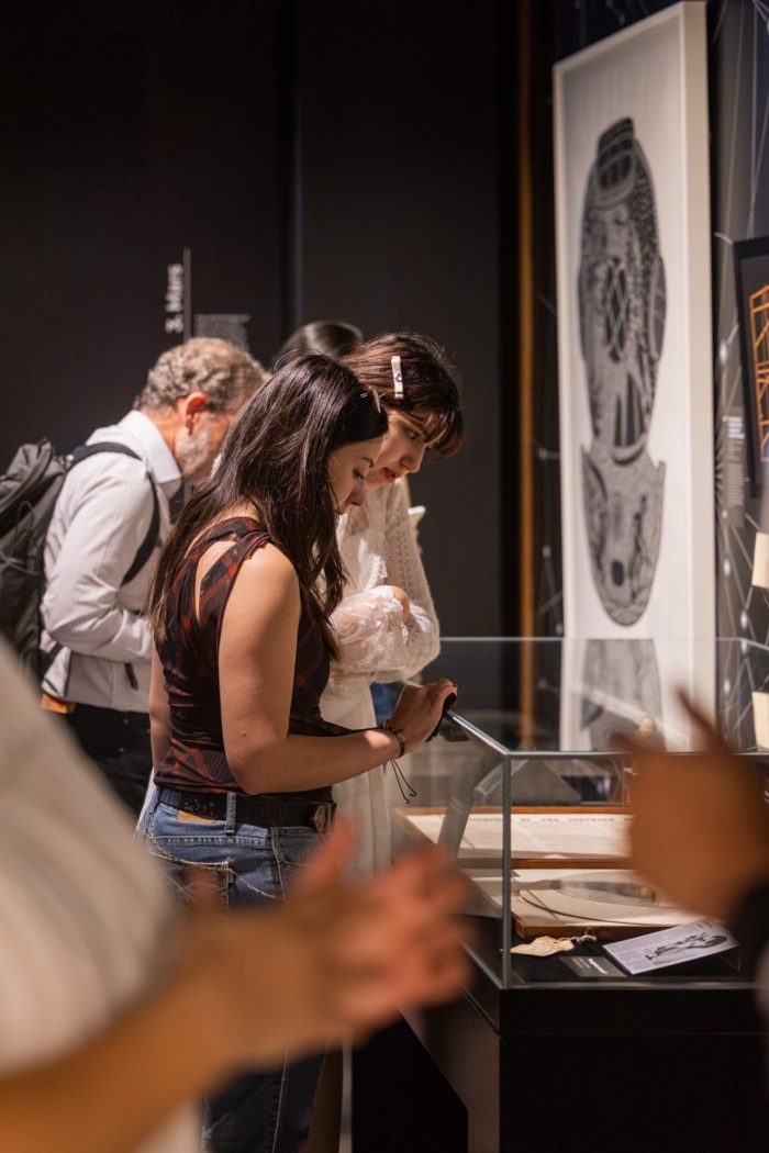 Two visitors looking closely at a glass display case containing documents and artifacts, with a large black and white artwork in the background.