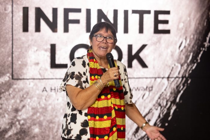 A woman wearing a vibrant Aboriginal flag-inspired scarf speaks into a microphone in front of an exhibition wall titled "Infinite Look”