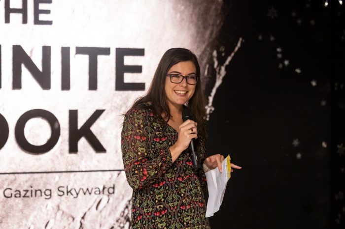 A woman in a floral dress smiling and speaking into a microphone in front of the "Infinite Look" exhibition title wall.