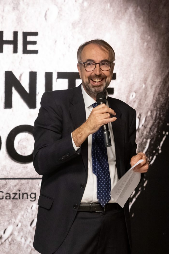A man in a suit and tie speaking into a microphone in front of "The Infinite Look" exhibition title.