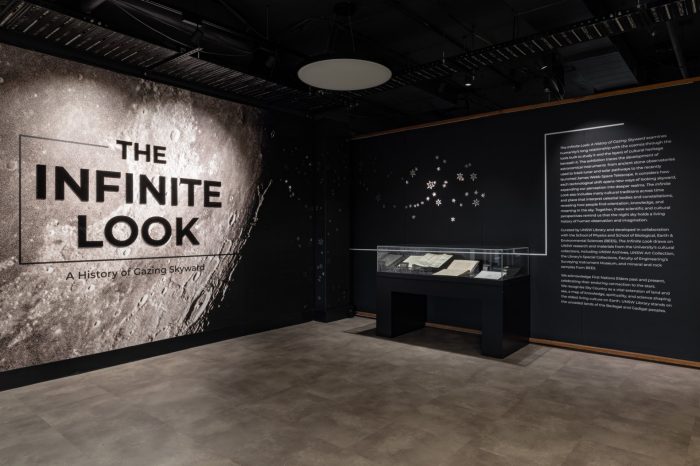 Exhibition title wall for "The Infinite Look" featuring a large moon image, an introductory text and a display case.