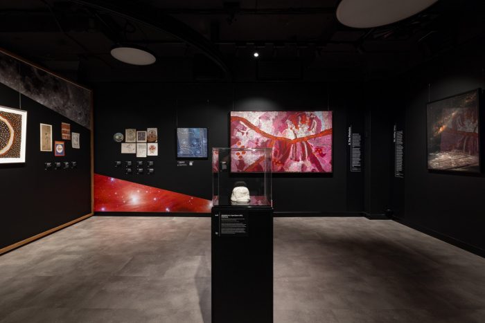 Exhibition gallery featuring Indigenous artworks on dark walls and a central display pedestal with a white sculpture in it.
