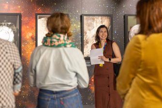A woman stands in a gallery space, addressing an audience. Behind her is several large framed images of the moon.