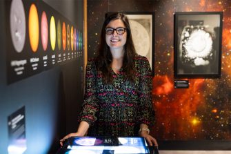A smiling woman stands in an exhibition space surrounded by colourful images of the sun, moon, and galaxies