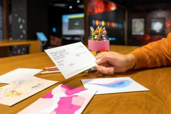 A hand holding a postcard with a message written on it. Various drawings and colour pencils surrounds it on the table.