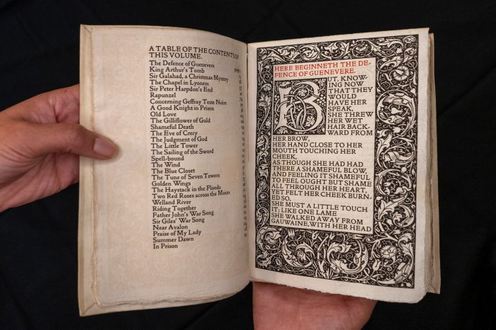 An open book is held in hands, displaying a table of contents on the left page and a page of text featuring a large, ornate initial letter and dense floral borders on the right.