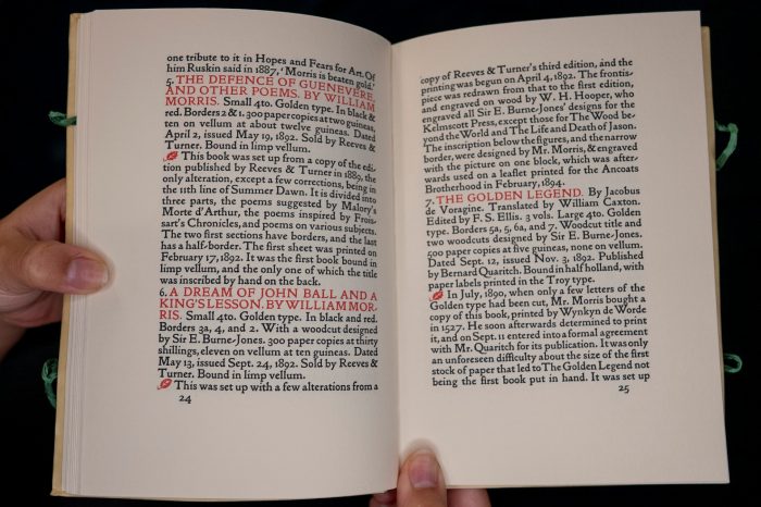 An open book held in hands displays two pages of dense, black typography, with occasional headings highlighted in red ink and small leaf-shaped decorative symbols marking the start of paragraphs.