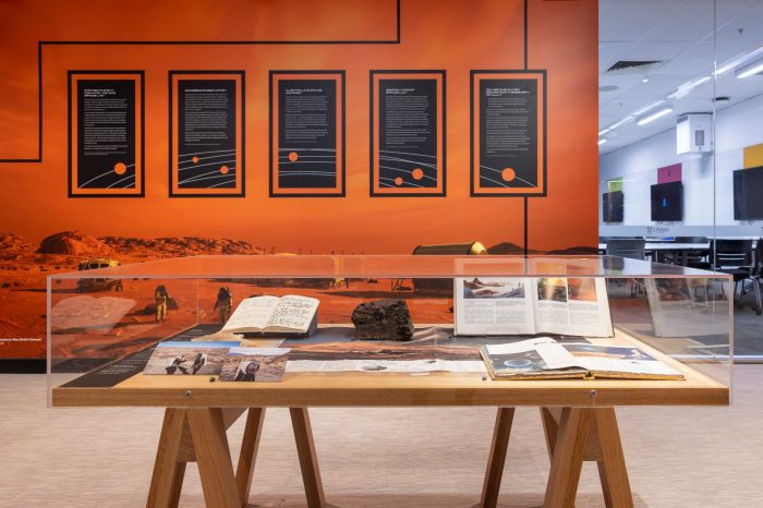 A wooden display table with a glass case containing open notebooks, books, photos, and a dark rock specimen, set against an orange exhibition wall with five black informational panels.