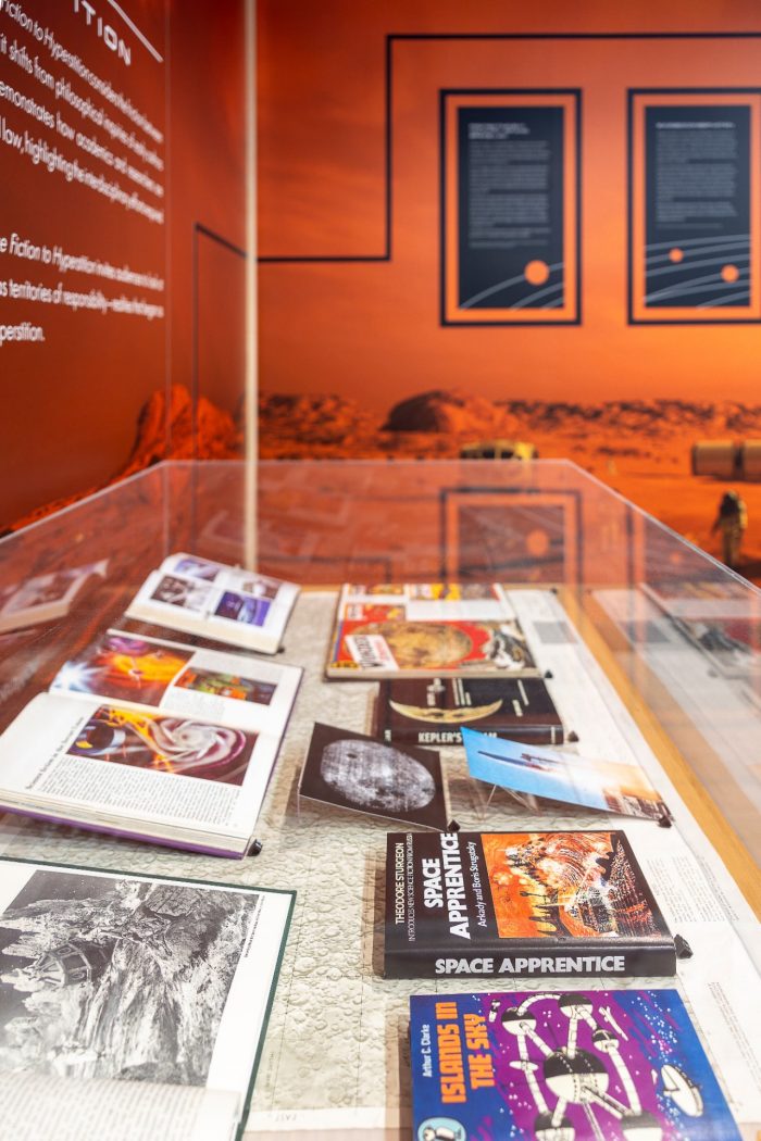 A high-angle view of a glass display case containing various science fiction books, including "Space Apprentice" and "Islands in the Sky", set against an orange exhibition wall.