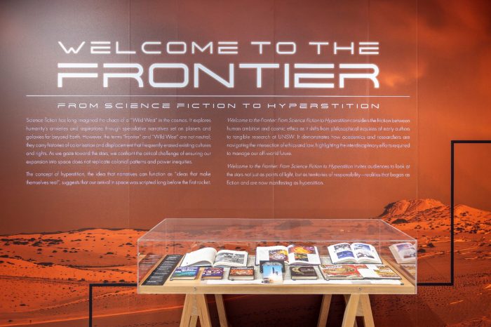 An exhibition wall with text reads "Welcome to the Frontier: From Science Fiction to Hyperstition", featuring a glass display case below it filled with books and magazines on a desert landscape background.