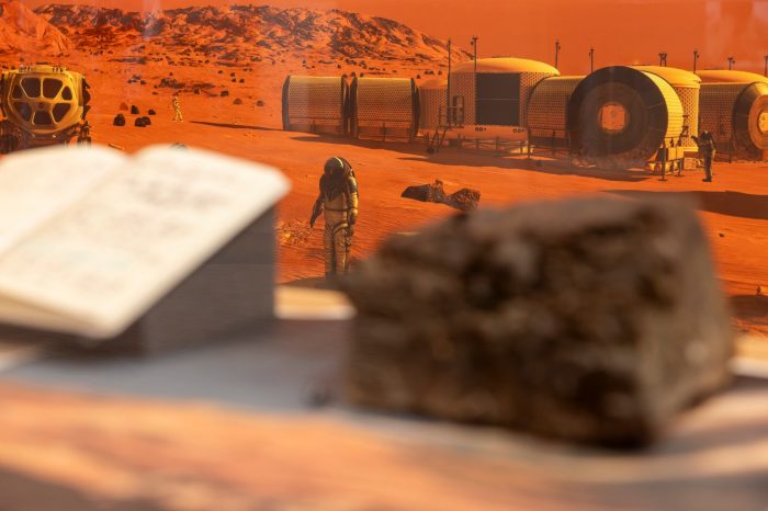 A close-up shot looking through a glass display case at a background illustration of an astronaut standing near a space habitat base.