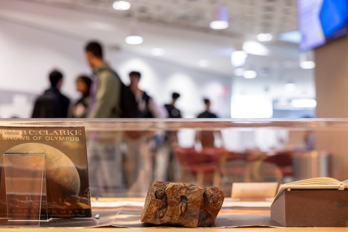 A museum display case containing a Martian science fiction book and a rock specimen, with a blurred crowd of people in the background.