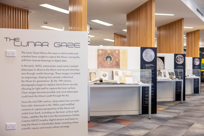 An exhibition display titled "The Lunar Gaze" with an informative text panel on the left, historical books and illustrations in display cases, and info panels and maps behind them.