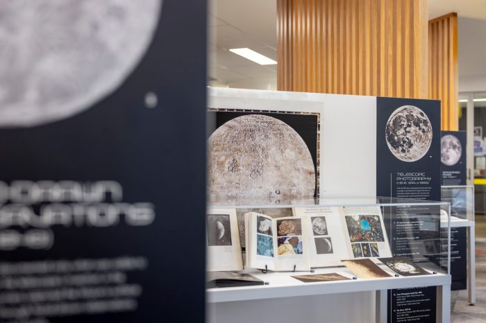 A glass display case in an exhibition showcasing open books and prints of historical telescopic photography of the moon.