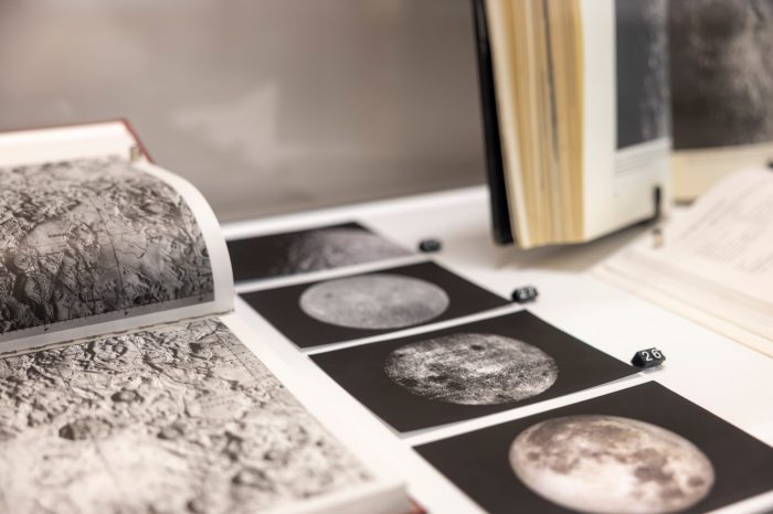 A display case with an open book displaying a textured lunar surface map alongside a row of small, black-and-white photographs tracking different phases and details of the moon.