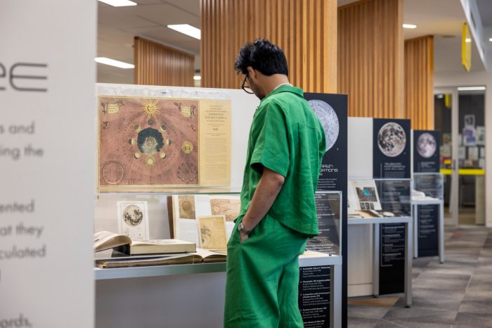 A man in a green matching short-sleeve shirt and trousers stands with his hands in his pockets, leaning forward to look at historical celestial illustrations in a display case.