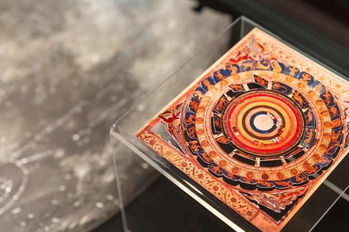 Close-up of a brightly coloured circular zodiac-style artwork displayed in a clear acrylic stand, with a large moon image blurred in the background inside an exhibition case.