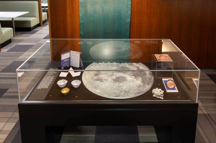 Glass display case featuring a large moon puzzle at the centre, surrounded by moon-themed artworks, folded paper pieces, small decorative objects, and exhibition labels against a wood-panelled backdrop. A long green flowing material hangs against the wall in the background. 