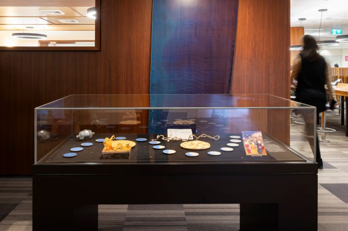 Glass display case containing moon-themed artworks and objects, including circular moon-like forms, illustrated cards, and small sculptures arranged in a lunar phase pattern, set against a wood-panelled wall. A long blue flowing fabric hangs against the wall, and a visitor is walking past in the background.