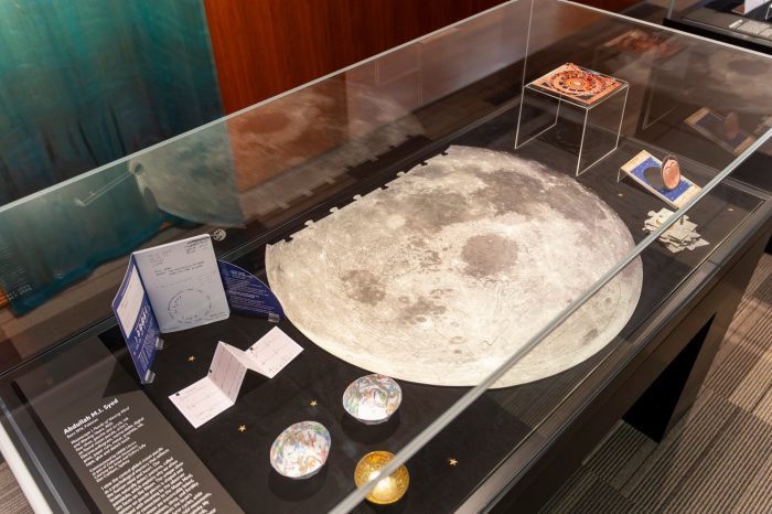 Glass display case featuring a large moon puzzle surrounded by small trinkets, folded paper pieces, and exhibition labels. 
