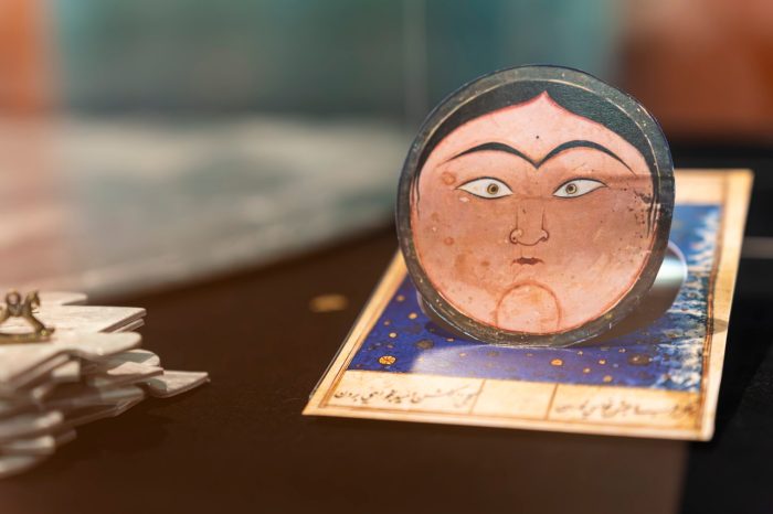 Close-up of a small circular illustration depicting a face on a moon-like surface on a starry blue card, displayed inside an exhibition display case.