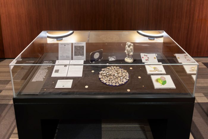 Glass display case containing moon-themed artworks, documents, silver coins arranged in a circular pattern, and an astronaut figurine against a wood-panelled backdrop.