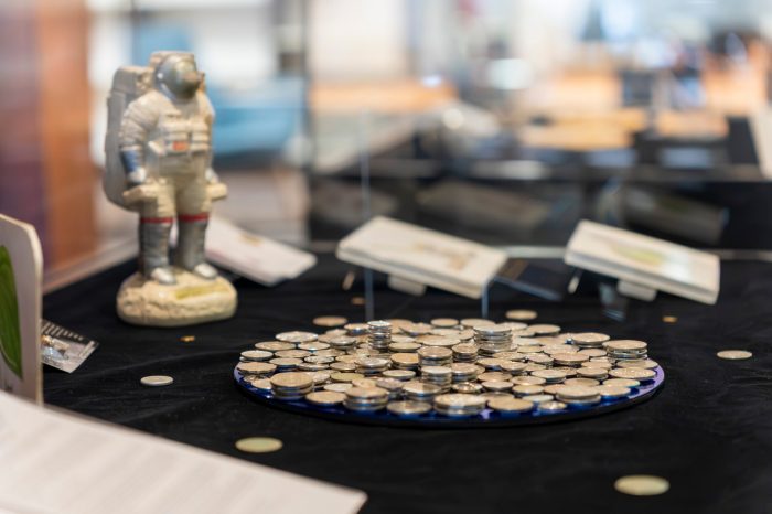 Close-up of the inside of a glass display case featuring a small astronaut figurine and a circular arrangement of stacked silver coins, and other objects blurred in the background.