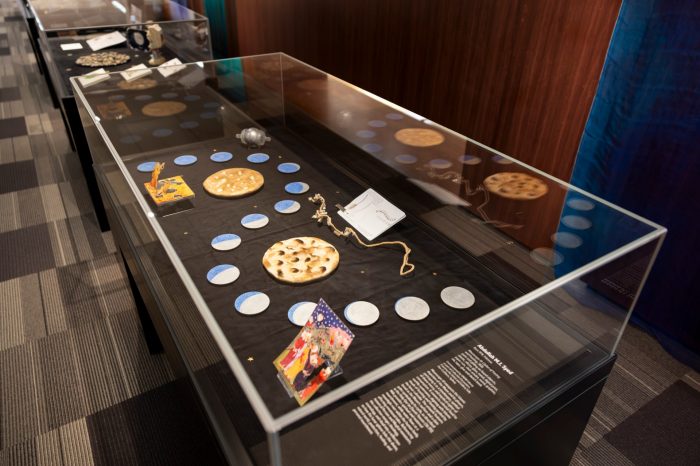 Long glass display case against a wood-panelled wall. Inside the display case is an assortment of moon-themed artworks and objects, including circular moon-like pieces, illustrated cards, and small sculptures.