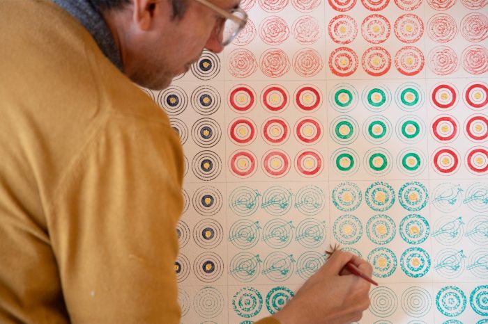 A man wearing a yellow sweater leans toward a large, grid-patterned artwork, meticulously applying gold paint to the centre of a green-coloured bird motif with a small brush.